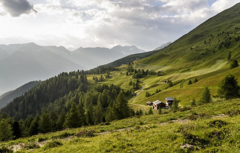 Paesaggio alpino soleggiato con baite sparse, boschi e montagne sotto un cielo parzialmente nuvoloso.